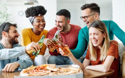 A group of young adults sitting around a table with a pizza while doing a toast with bottles of beer.