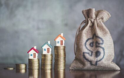 five progressively taller stacks of coins with mini houses on top leading to a canvas sack with a dollar sign on it.