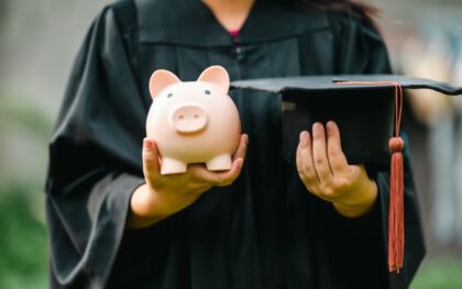 Closeup of a high school graduate in a black gown holding a graduation cap and a piggy bank to save money for college.