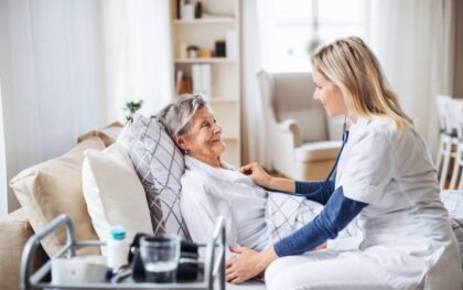 A senior patient lies on a white sofa in their living room while a home healthcare worker examines them with a stethoscope.