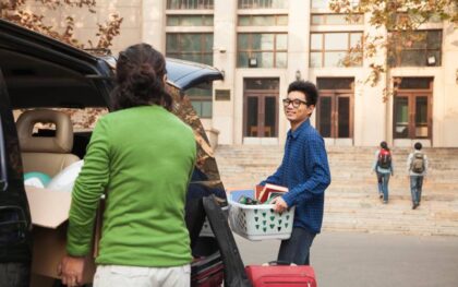 A dorm building's exterior. A young man holding a bin of school supplies smiles at his mom who is unloading the car.
