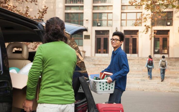 A dorm building's exterior. A young man holding a bin of school supplies smiles at his mom who is unloading the car.