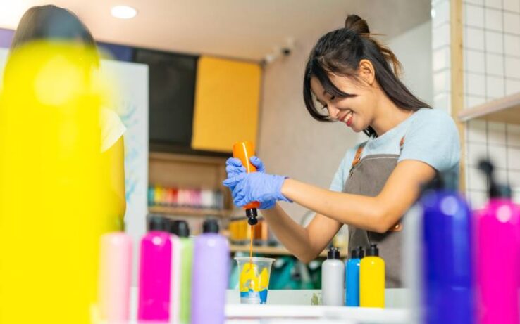 A young woman in a small art studio squeezing orange paint into a plastic cup with other liquid paints inside it.