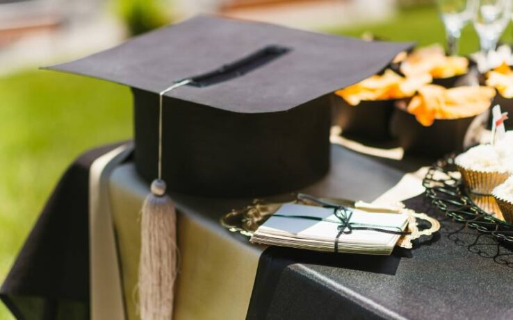 A black table with a graduation cap, a bundle of cards sitting on a gold platter, and snacks and glasses.