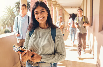 A young college student smiling as she holds her books and walks down an outdoor corridor on her college campus.