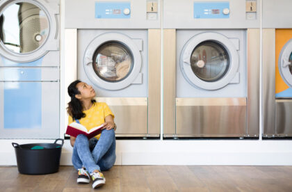 A woman sits in front of a washing machine at a laundromat. She has an open book in her lap and a laundry basket beside her.