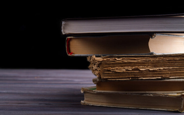 A staggered stack of new and old hardcover books on a table in front of a black background. The books' pages face the viewer.