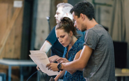 Three college students working together on a theater set. One student gestures to the script while explaining something.
