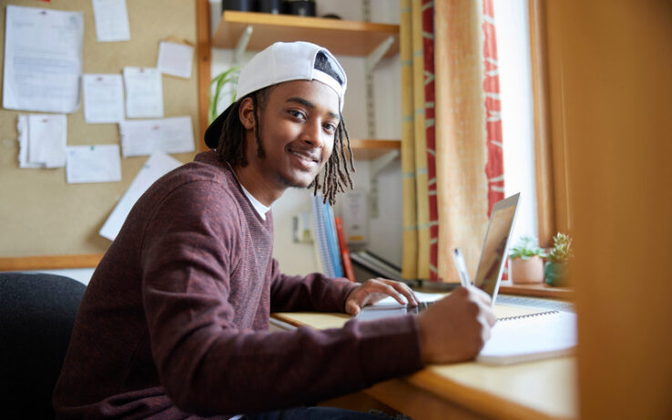 A young man wearing a backward baseball hat holding a pen at a wooden desk with an open laptop and notebook facing a window.