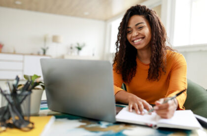 A young college student smiling at her laptop while sitting in her room and taking notes in a notebook.