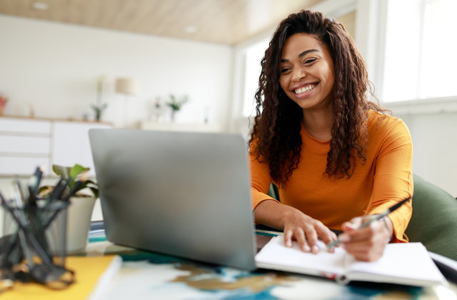 A young college student smiling at her laptop while sitting in her room and taking notes in a notebook.