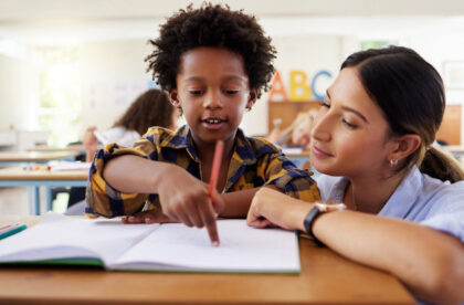 A young woman in a blue button-down shirt crouches beside a desk where a young boy points at a paper.