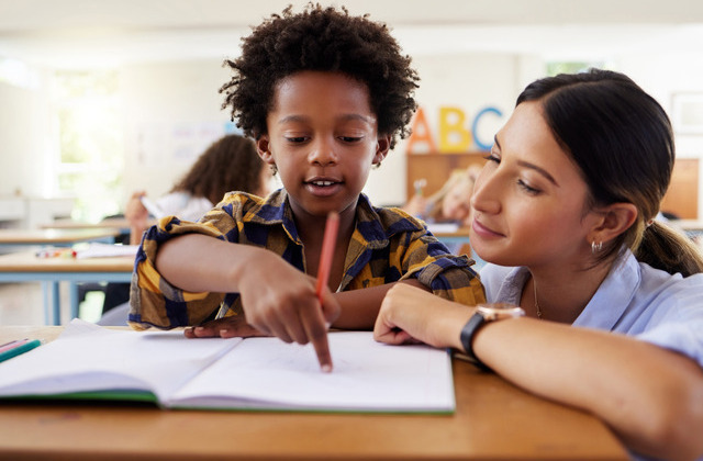 A young woman in a blue button-down shirt crouches beside a desk where a young boy points at a paper.