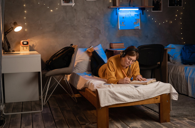 A young woman wearing headphones and writing in a notebook on a twin bed in a small room decorated with fairy lights.