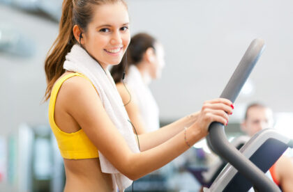 A young woman smiles while she walks on a treadmill and listens to music with her earbuds. Her hands grip the handlebars.