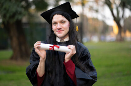 A young woman dressed in a black cap and gown smiling as she hold a rolled diploma tied with a red ribbon between her hands.