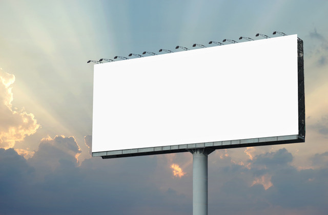 A large billboard displays a blank, white background. The sun shines through the clouds behind the billboard.