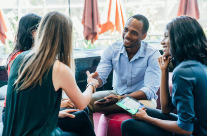 A group of professional-looking individuals are sitting in a circle. One woman and a man are shaking hands and smiling.