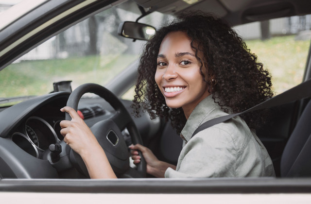 A young woman sitting in a sedan's driver's seat with her hands on the steering wheel while smiling.