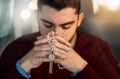 A religious individual with their hands folded together as he prays with a rosary. The rosary is woven into his fingers.