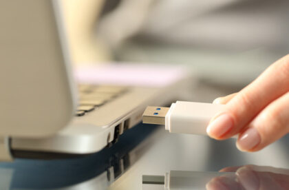 A close-up view shows a person holding a white flash drive next to an open silver laptop. The background is blurry.