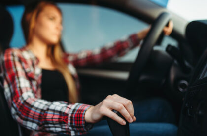 A young woman in a red flannel shirt driving a vehicle with one hand on the gear stick and the other on the wheel.