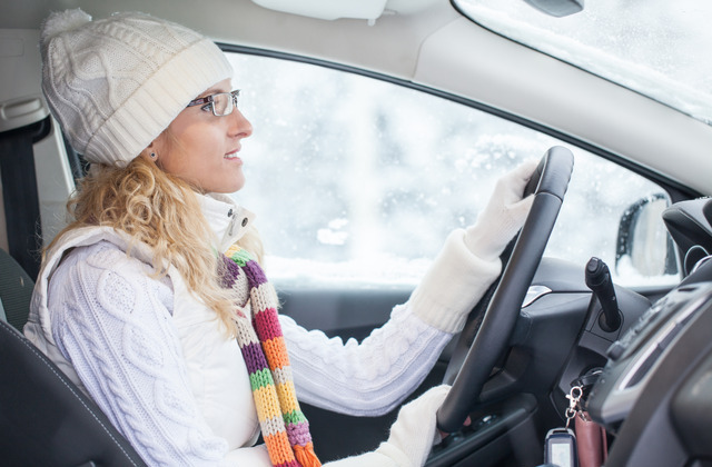 A woman in winter attire sitting behind the wheel of her vehicle. She is currently driving through snowy conditions