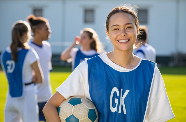 A smiling girl wears a blue jersey that reads "GK" and holds a soccer ball. Four players stand in a group in the distance.