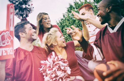 College students in school spirit gear cheering. One is holding a pom-pom and another is holding a foam finger.