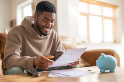 A smiling Black man sitting on a couch and reviewing paperwork at a table with a small blue piggy bank to his left.