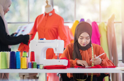 A young woman sitting at a table with many spools of colorful thread, a white sewing machine, scissors, and other sewing tools.