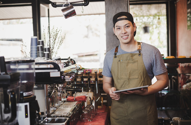 A young man in an apron standing in front of a professional espresso machine with two portafilters in a coffee shop.