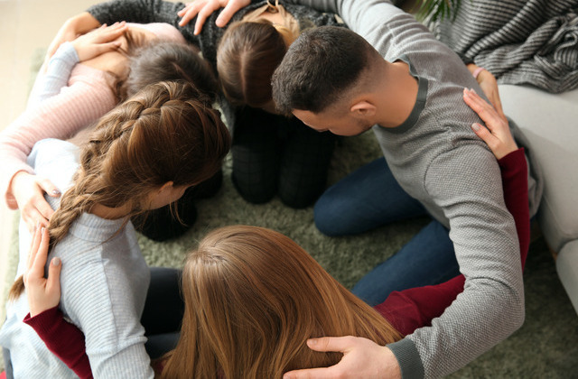 Young people huddle together in a circle with their arms around each other. They are bowing their heads.