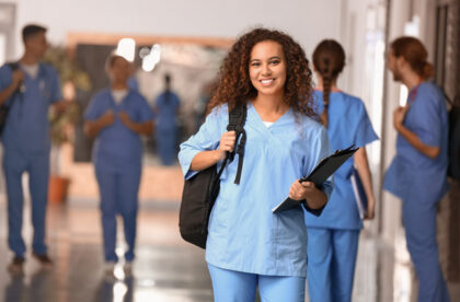 A medical student in blue scrubs holds a black backpack over her right shoulder and a black clipboard in her left hand.