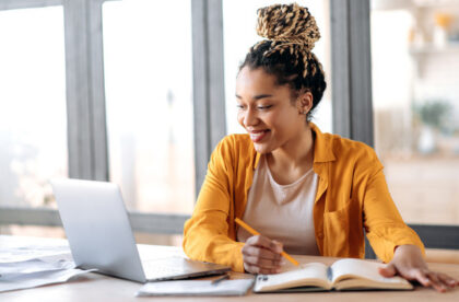 A young woman sits at a desk and smiles while looking at a laptop. She writes in a notebook with a pen.