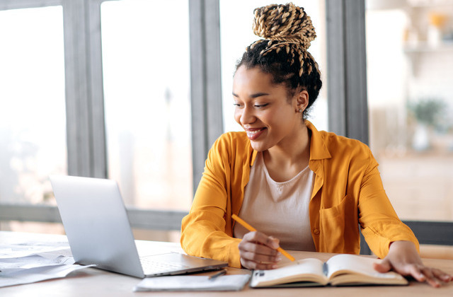 A young woman sits at a desk and smiles while looking at a laptop. She writes in a notebook with a pen.