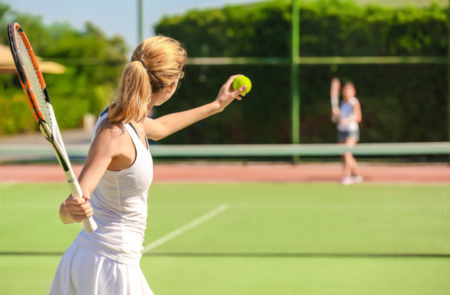 Two women stand on a tennis court. One woman prepares to serve the ball while the other waits in the distance.