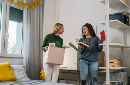 Two young women are laughing together in their dorm room. One is holding a box and the other has two books in her hands.