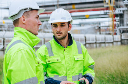 Two young engineers in white hard hats and high-visibility jackets talking while working at an oil and gas refinery.