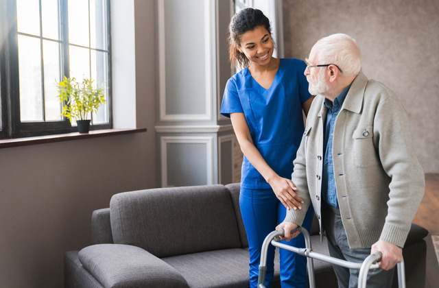 A young nurse in blue scrubs helps an elderly man stand. The man uses a walker and wears a tan jacket.