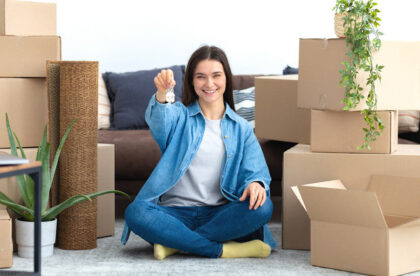 A young woman sits cross-legged in the living room of her new apartment surrounded by cardboard boxes holding a set of keys.