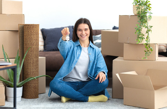 A young woman sits cross-legged in the living room of her new apartment surrounded by cardboard boxes holding a set of keys.
