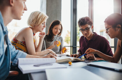 A racially diverse group of three women and two men at a table by a window with books and crumpled yellow sticky notes.
