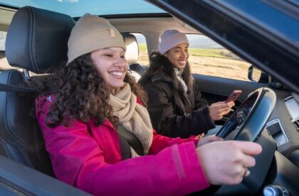 Two young college-aged women sit in a car smiling together. One sits in the passenger seat while the other drives the car.
