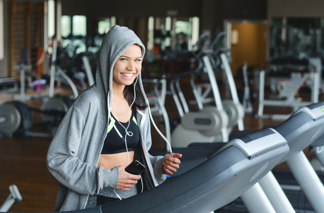 A young woman smiling while running on a treadmill in a gym. She's wearing a hoodie over her head and holding her phone.