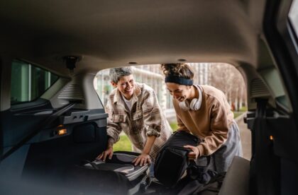 One younger and one older woman smile at each other as they load luggage into the trunk of a large vehicle.
