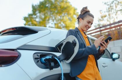 A smiling young woman looking at her smartphone and leaning against a white electric vehicle as it charges.