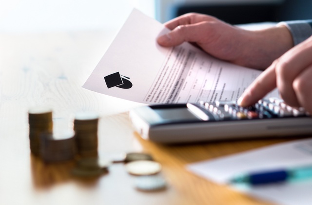 Coins sit on a table while a person uses a calculator and holds a document with a graduation cap on it in their hand.