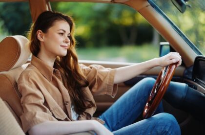 A young woman sitting in her car with one hand on the wheel and the other resting on the arm rest in the middle.
