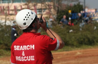 A man wearing a white hard hat and a red shirt with "search and rescue" on the back speaks into a walkie-talkie.